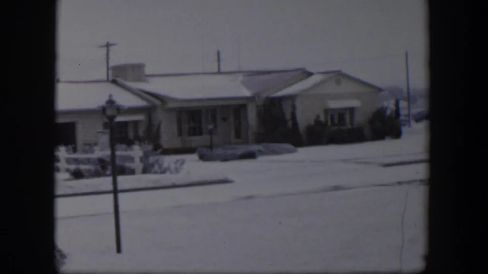 1954 Nevada winter frame with a ranch-style home, snow-covered roof and lawn, foreground lamp post, and utility poles.