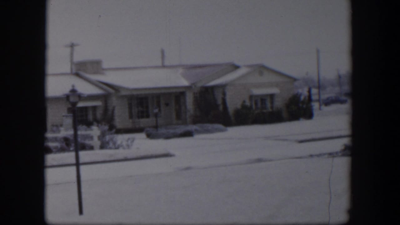 Snow-covered single-story house across a quiet residential street with a lamppost in front.