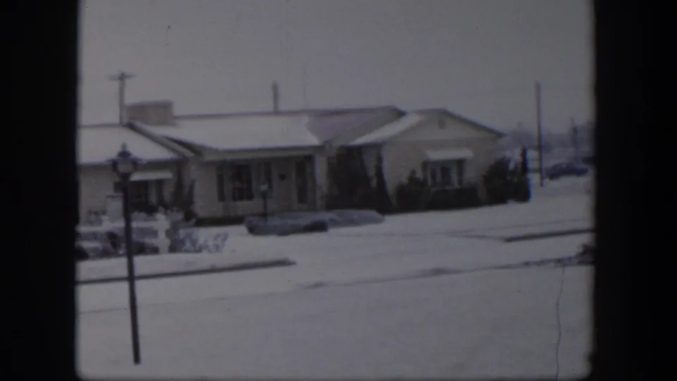 1954 Nevada home-movie frame showing a snow-covered single-story house, front yard, street, and lamppost.