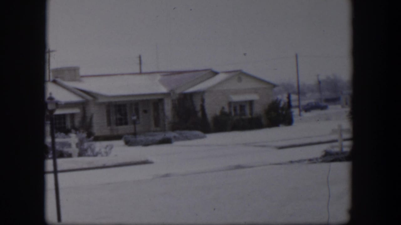Snowy residential street with a low house, shrubs, utility poles, and a parked car in the distance.