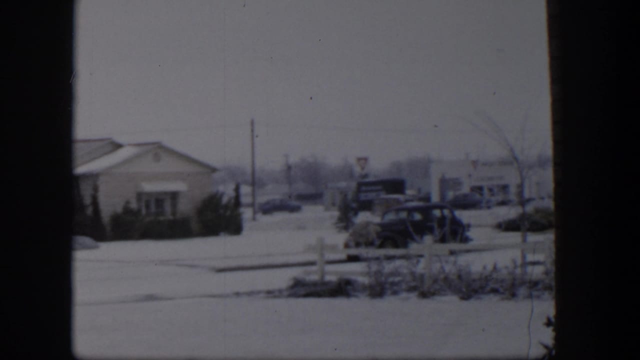 1954 Nevada winter street view with a low building, snow-covered ground, older parked cars, and roadside signs seen through an 8mm frame.