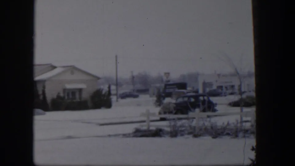 1954 Nevada winter street view with a low building, snow-covered ground, older parked cars, and roadside signs seen through an 8mm frame.