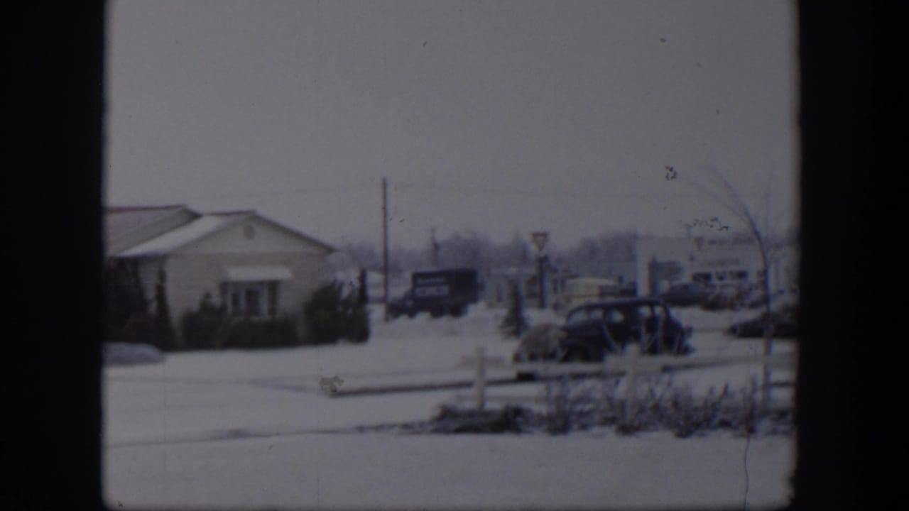 Snowy roadside scene with low building, parked dark car, signs, and distant traffic.