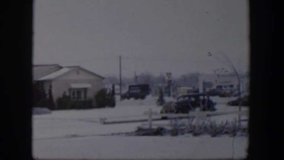 A 1954 Nevada street in winter, showing a low building, snow on the ground, a dark parked car, roadside signs, and distant vehicles.