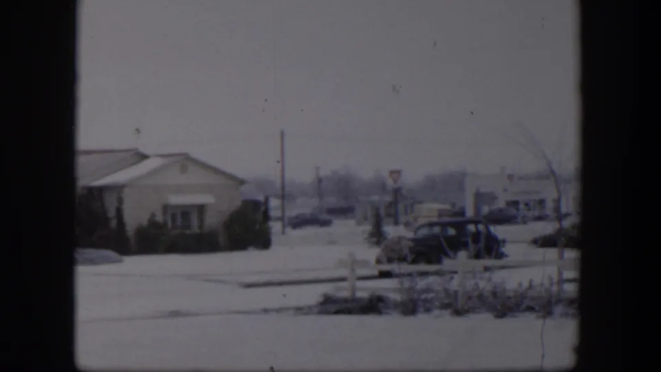 Snowy 1954 Nevada intersection with a low roadside building, older automobiles, signposts, shrubs, and muted 8mm home-movie grain.