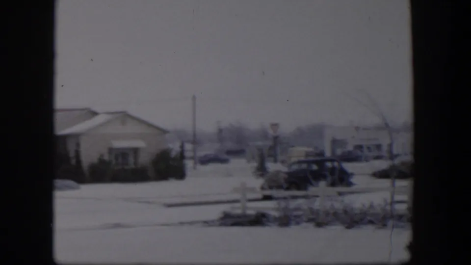 Final 1954 Nevada winter frame showing a low building, snowy roadside, parked car, signposts, and distant traffic under a gray sky.