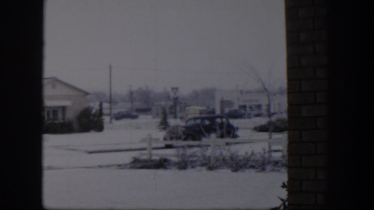 Snowy suburban street with a parked dark car, low houses, and dark window framing.