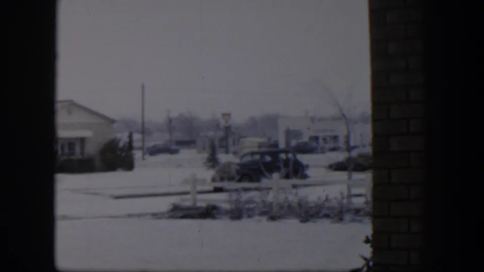 1954 Nevada home-movie frame showing a snow-covered residential street, a parked car, distant houses, shrubs, and dark interior framing.