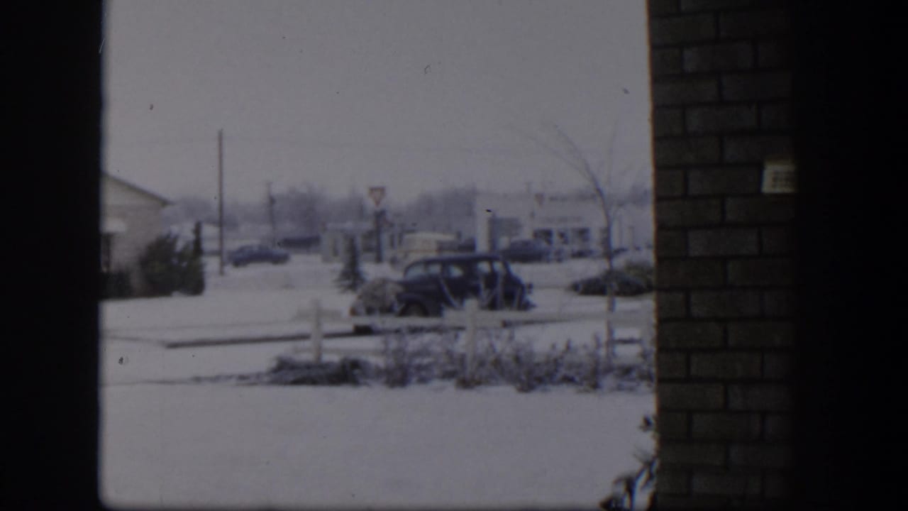 Snowy residential street seen through dark framing, with parked cars and houses beyond.