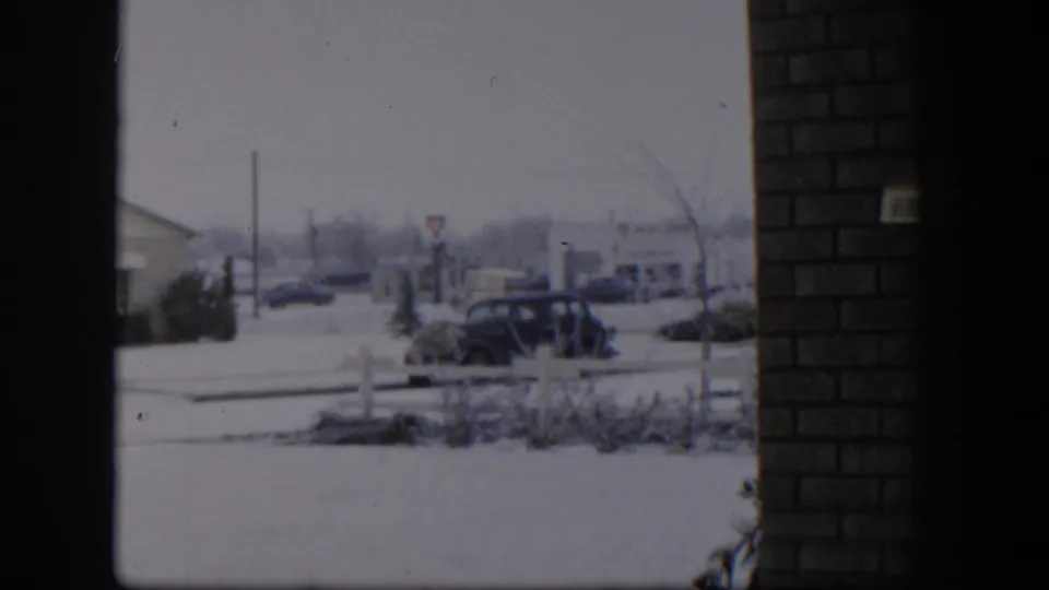 1954 Nevada frame of a snow-covered suburban street, parked cars, low homes, shrubs, and a brick edge at the right side of the view.
