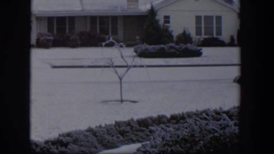 1954 Nevada home-movie view across a snowy lawn and street toward a low suburban house with front shrubs and a young bare tree.