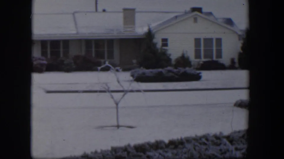 1954 Nevada home-movie frame showing a snow-dusted suburban yard, bare young tree, shrubs, and a single-story house beyond the lawn.
