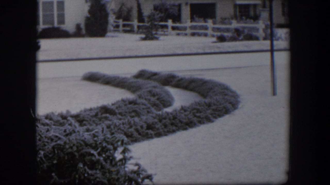 1954 Nevada 8mm frame showing a snowy suburban street, curved hedge, white fence, and single-story bungalow.