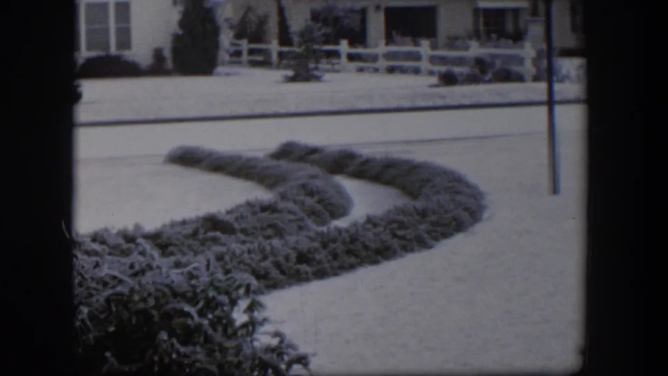 1954 Nevada 8mm frame showing a snowy suburban street, curved hedge, white fence, and single-story bungalow.