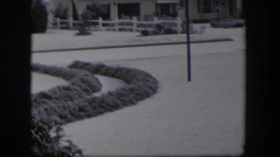 1954 Nevada home-movie frame with snow on the street, hedge, fence, and a low suburban bungalow.