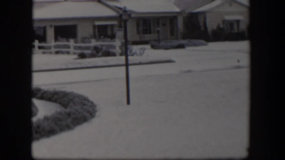 1954 Nevada 8mm view of a snowy bungalow frontage with fence, street, hedge, and utility pole.