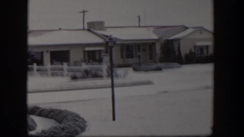 1954 Nevada frame showing a snow-covered residential bungalow, white fence, utility pole, and quiet street.