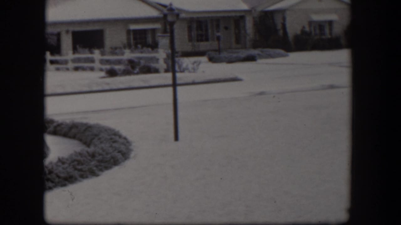 Snowy residential street with a house, fence, and central pole.