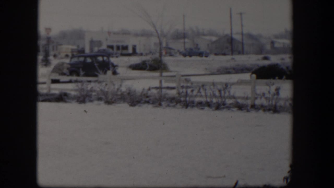 Snowy street seen through dark window framing, with fence, shrubs, cars, and low buildings.