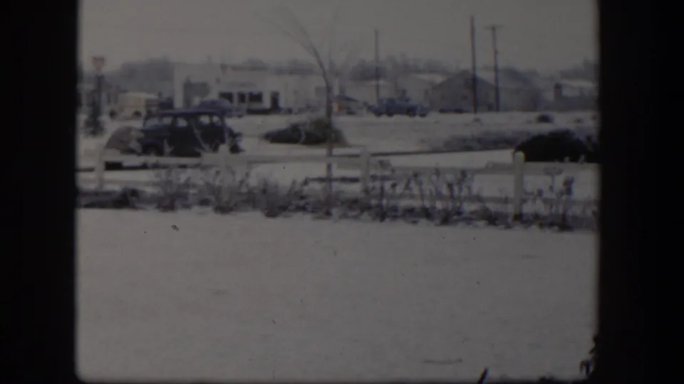 1954 Nevada winter street view through a window, showing snow, a white fence, shrubs, parked cars, and low buildings.
