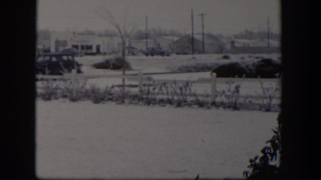 Snow-covered roadside with dark window divider, parked cars, fence, shrubs, and distant buildings.