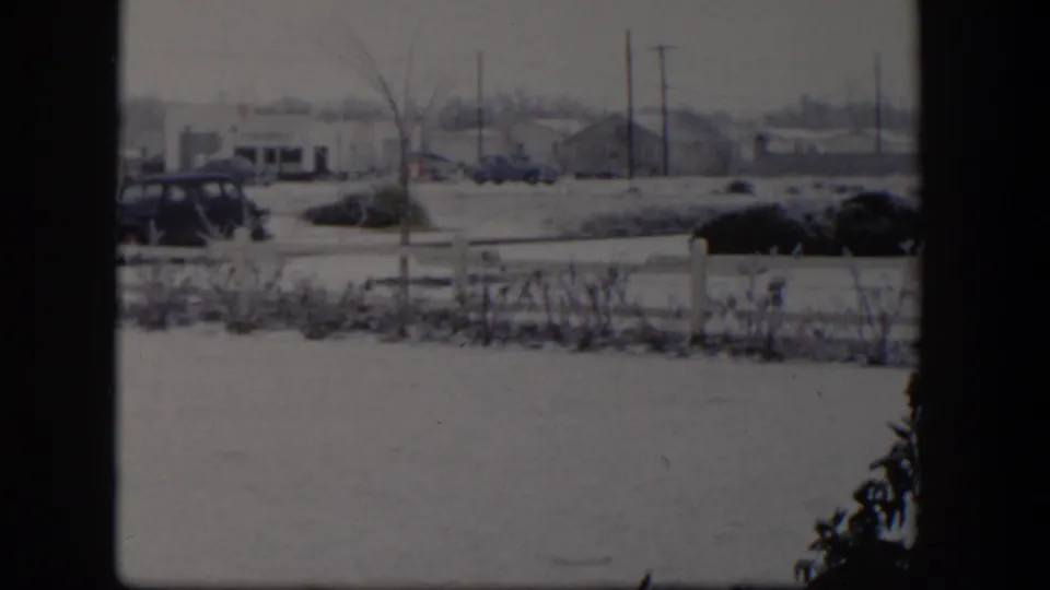 1954 Nevada snowy roadside scene framed by a dark window divider, with cars, fence, shrubs, utility poles, and low buildings.