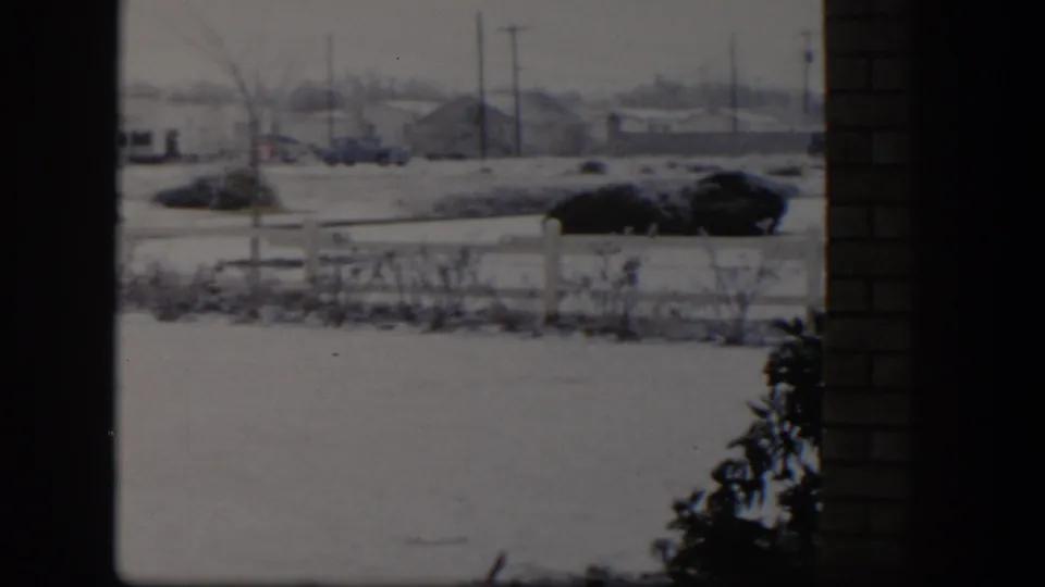 1954 Nevada winter view with a snowy foreground, shrubs, fence, dark car, distant buildings, and a brick edge at right.