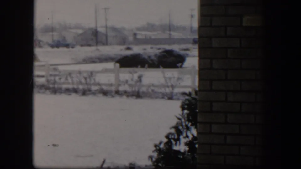 1954 Nevada snowy street view through dark framing and brickwork, showing fence, shrubs, a dark car, utility poles, and distant buildings.