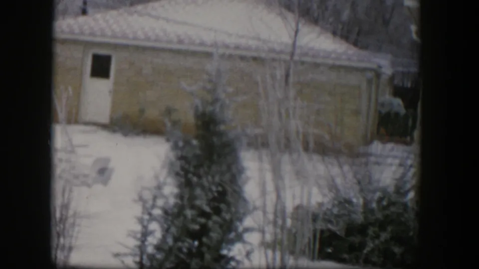 1954 Nevada 8mm frame with a yellow residential wall, white door, snowy yard, bare shrubs, and a small evergreen tree.