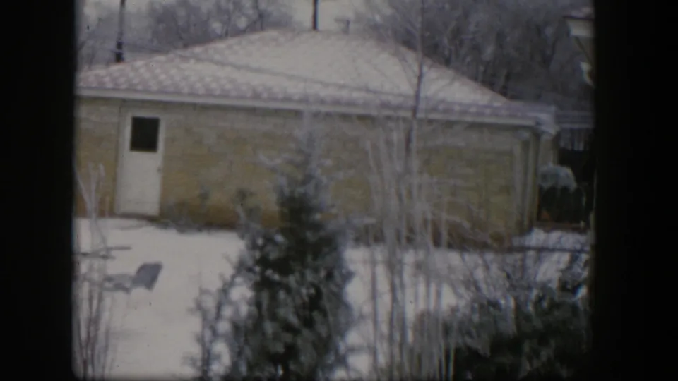 1954 Nevada home-movie frame showing a snow-covered suburban yard, single-story house, pale side door, evergreen tree, and bare winter branches.