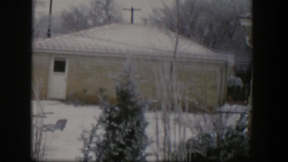 1954 Nevada frame of a quiet snowy yard with a low residential building, side door, evergreen, bare trees, shrubs, and dark window framing.