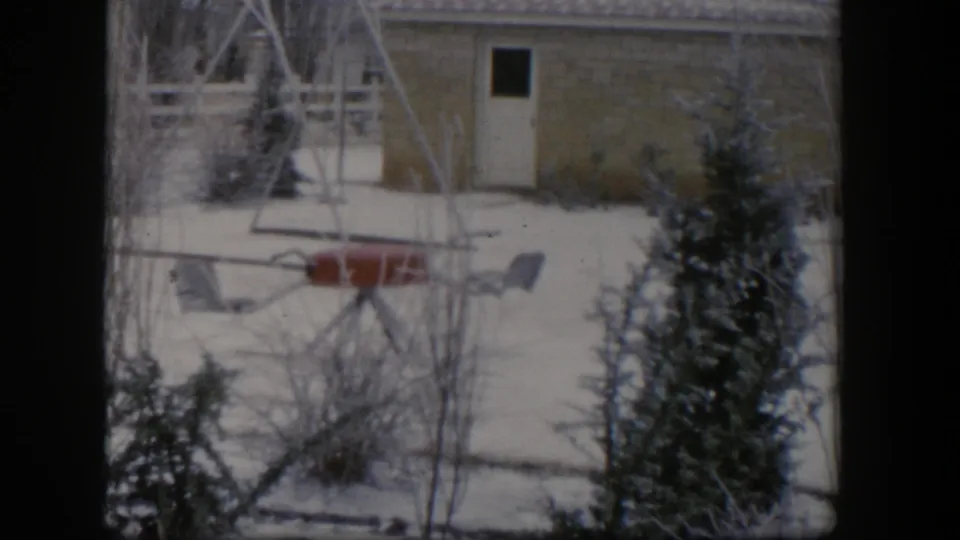 1954 Nevada 8mm still of a snowy backyard, with red playground equipment framed by bare trees, evergreen branches, and a house wall.
