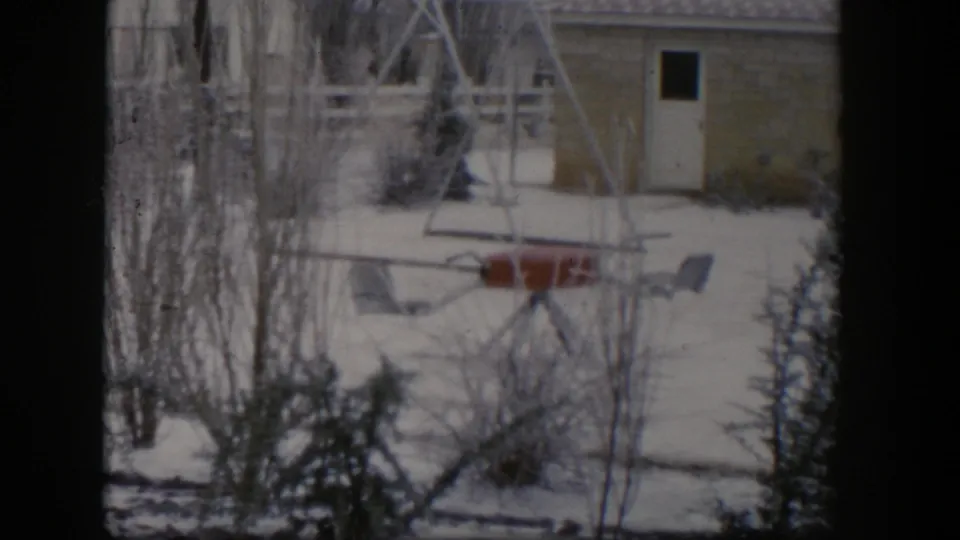 1954 Nevada home-movie frame showing a snowy backyard playset near bare branches, an evergreen tree, a brick wall, and a white door.