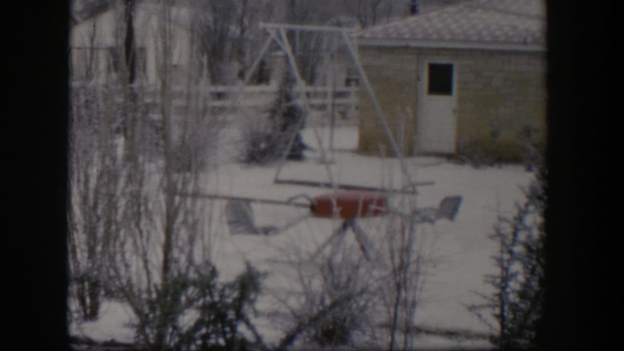 1954 Nevada 8mm frame showing a snow-covered residential yard with bare trees, a swing set, outdoor chairs, and a small shed.