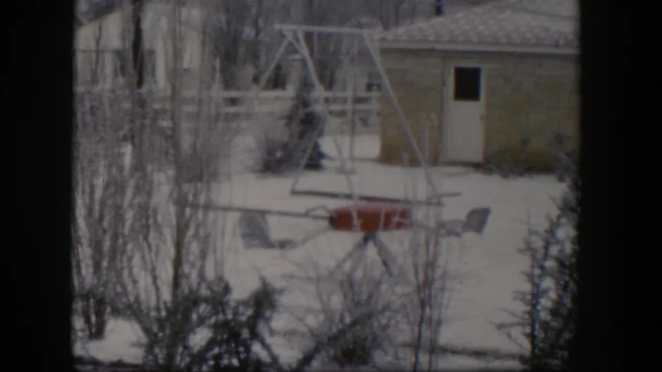 1954 Nevada 8mm frame showing a snow-covered residential yard with bare trees, a swing set, outdoor chairs, and a small shed.