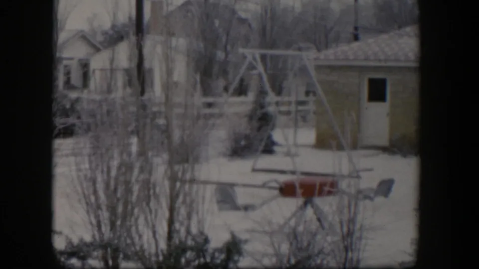 1954 Nevada home-movie frame of a snowy backyard, with a swing set and chairs framed by bare trees and nearby outbuildings.