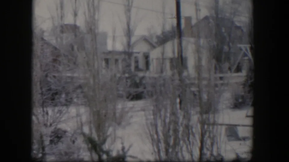 1954 Nevada 8mm frame showing a snow-covered residential yard with bare trees, small buildings, and the edge of a swing set.