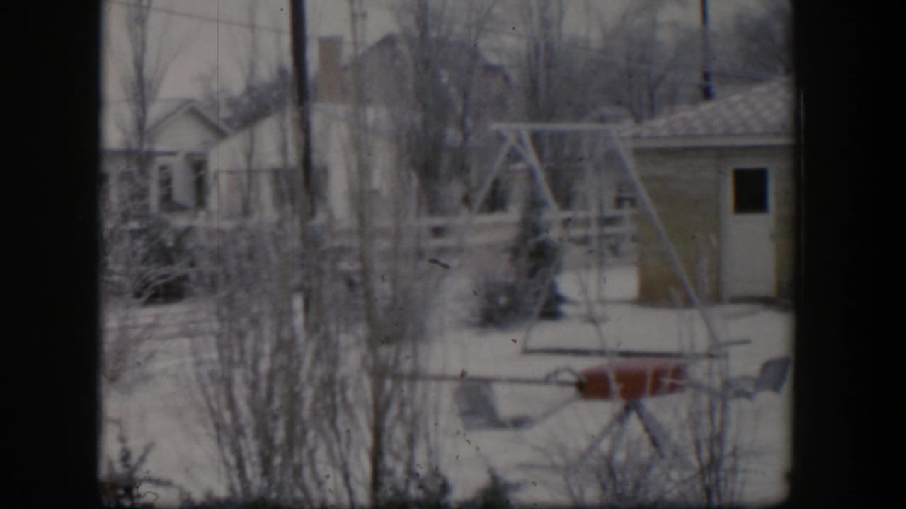 Snowy yard with swing set, red seesaw, bare trees, and a small outbuilding or garage.