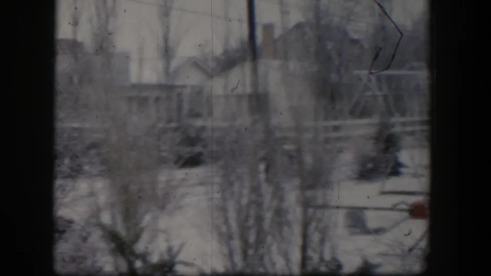 Grainy 1954 Nevada 8mm frame showing a snow-covered suburban yard, bare trees, background houses, and partial play equipment.
