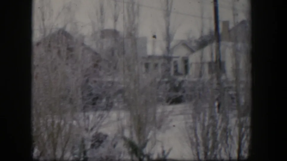 1954 Nevada 8mm frame of a snowy suburban backyard with bare trees, background houses, utility poles, and a partly visible play set.