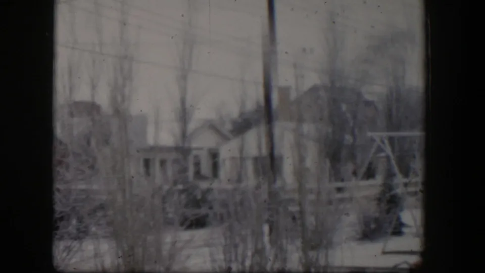 1954 Nevada home-movie frame with snow, leafless trees, a modest building, and a wooden frame structure at the right edge.