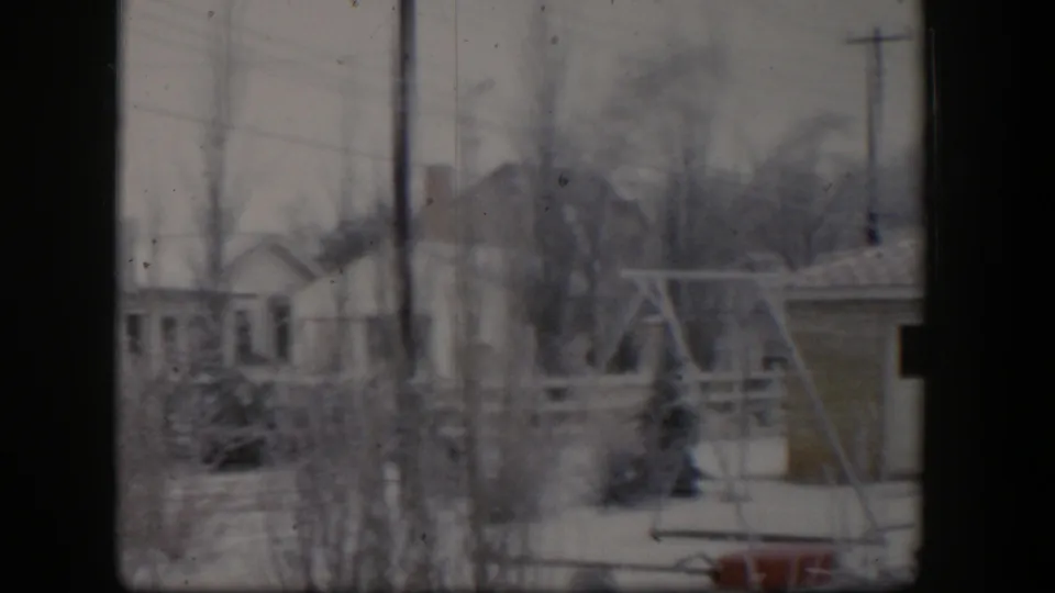 1954 Nevada 8mm frame showing a snow-covered backyard with a swing set, fences, bare shrubs, utility poles, and neighboring houses.