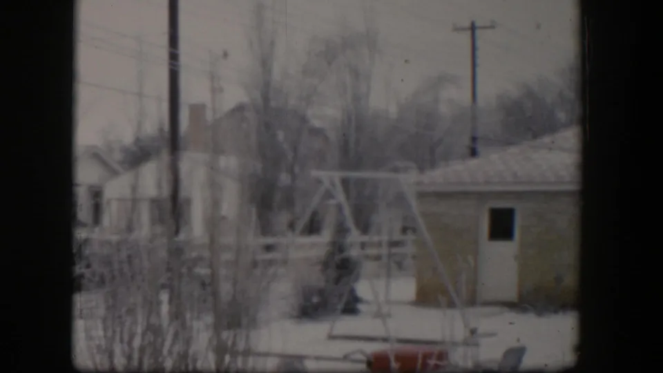 1954 Nevada home-movie frame of a snowy backyard with a swing set, small outbuilding, bare trees, utility poles, and nearby houses.