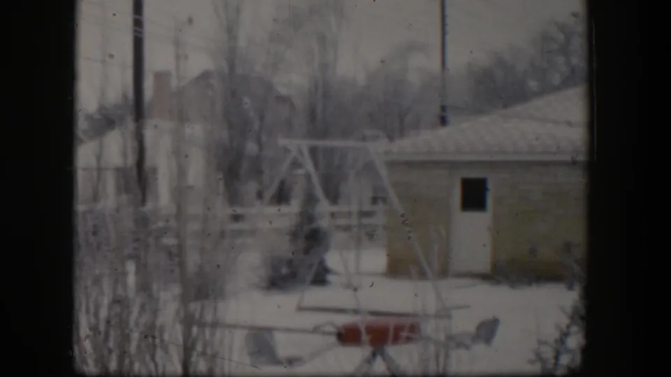 1954 Nevada 8mm view of a snow-covered residential yard with a swing set, small building, shrubs, fences, and rooftops beyond.