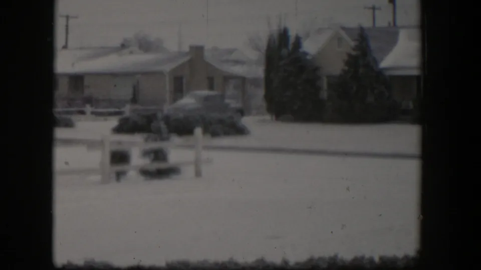 1954 Nevada frame showing snow around houses, a parked car, fences, shrubs, and utility lines in a residential area.