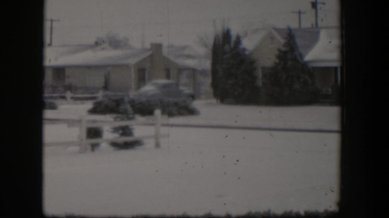 1954 Nevada home-movie frame showing a snowy residential street with a single-story house, parked car, fence, shrubs, and utility poles.