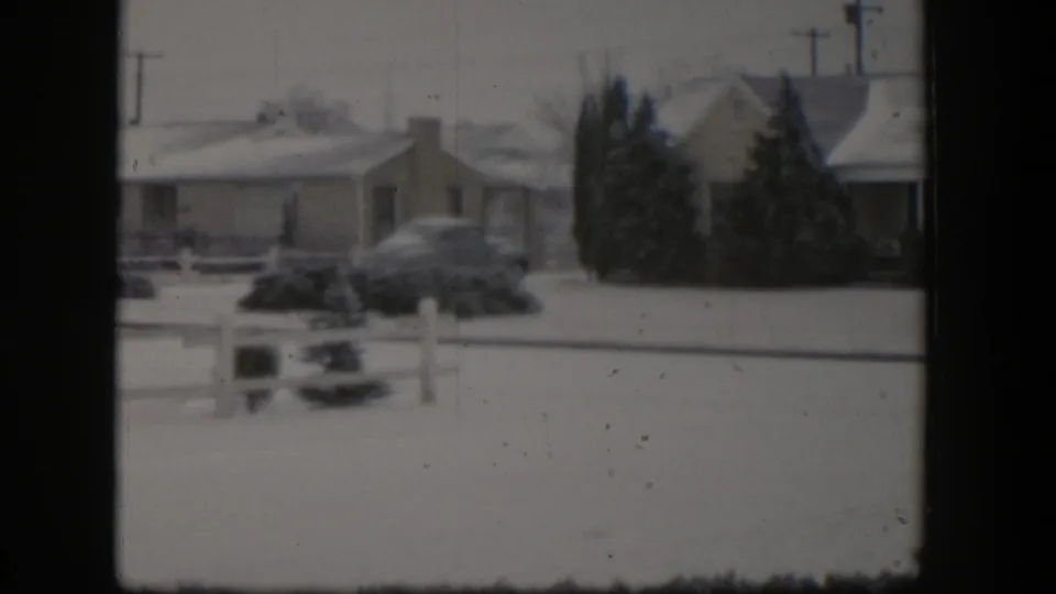 1954 Nevada home-movie frame showing a snowy residential street with a single-story house, parked car, fence, shrubs, and utility poles.