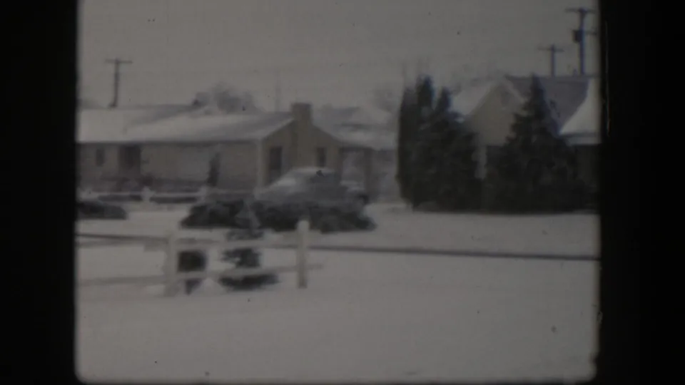 1954 Nevada frame of a snow-covered suburban house with a rounded parked car, low fence, dark evergreens, and utility poles behind it.