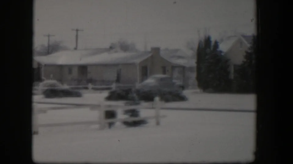 1954 Nevada winter frame with a low residential house, snow-covered yard, white fence rails, evergreen plantings, and a parked automobile.
