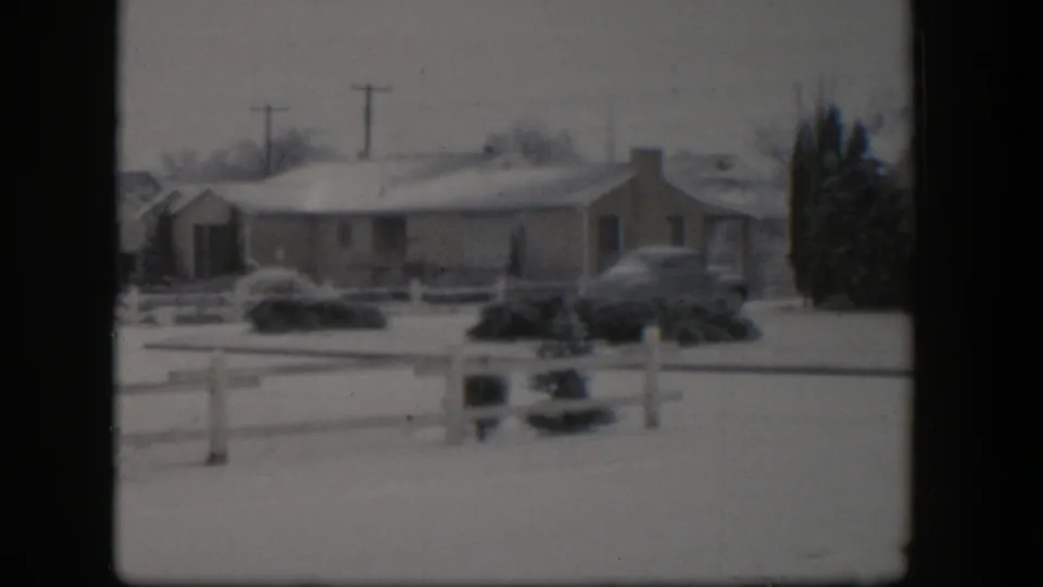 1954 Nevada home-movie frame showing a wider snowy suburban view with low houses, parked cars, fence rails, evergreen trees, and utility poles.